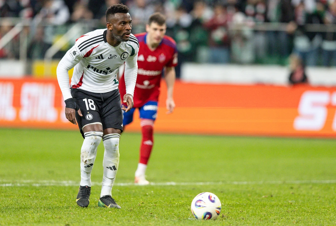 piłkarz w białym stroju z numerem 18 przygotowuje się do wykonania rzutu wolnego na boisku, w tle przeciwnik w czerwonym stroju oraz rozmyta publiczność na trybunach