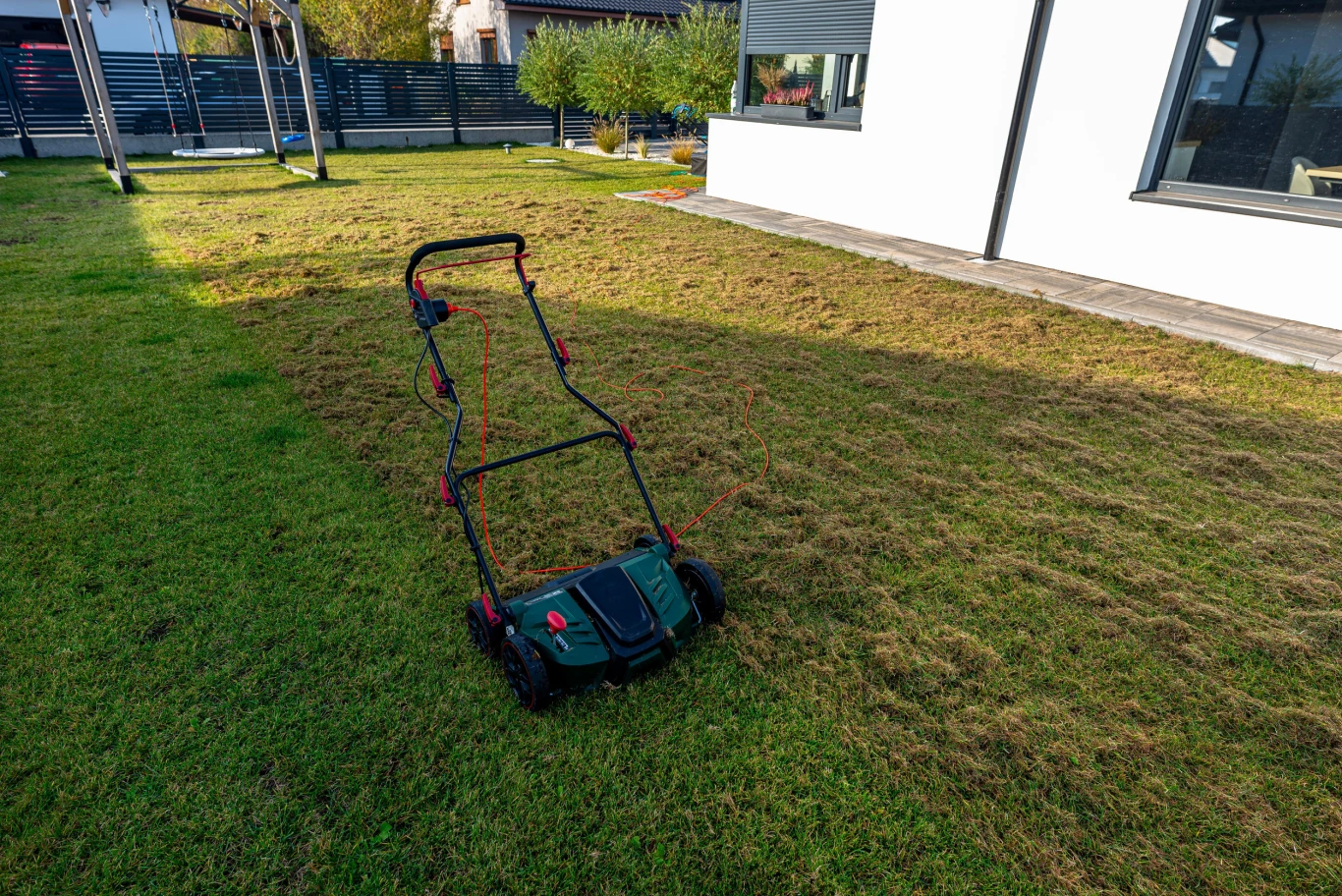 Kiedy wertykulować trawnik? Dobór terminu ma ogromne znaczenie. Elektryczny wertykulator na trawniku z widocznymi pasami napowietrzenia, w tle taras i ogrodzenie ogrodu