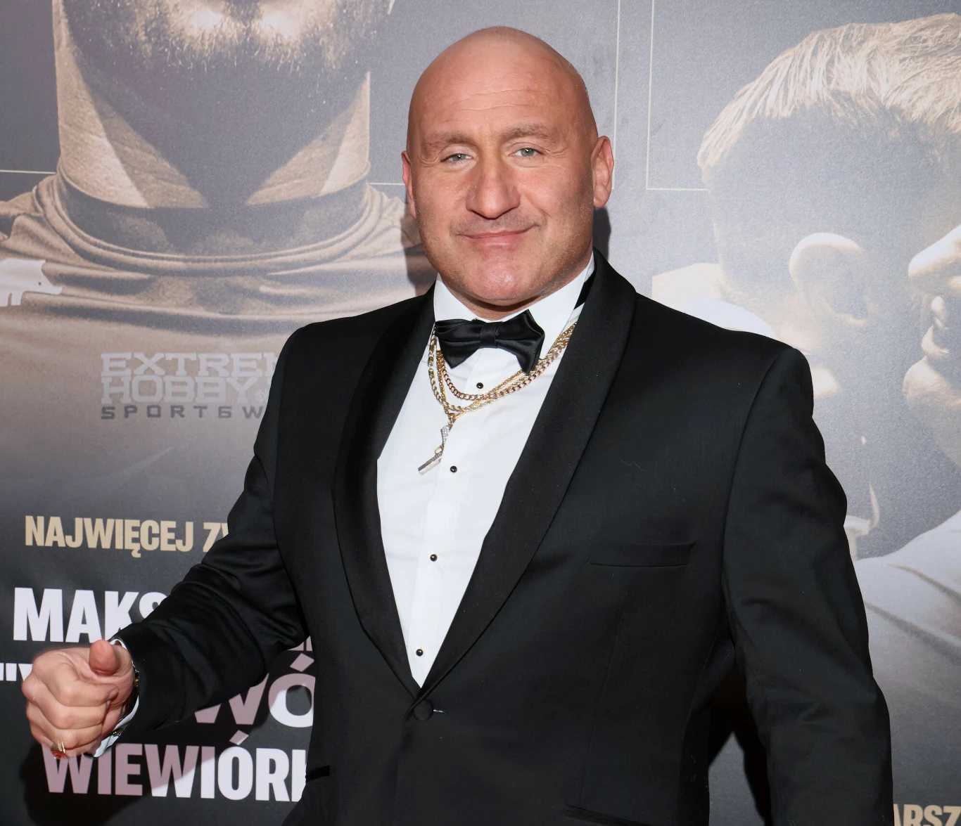 łysy mężczyzna w eleganckim smokingu z muchą i złotymi łańcuchami na szyi pozuje na tle ściany z grafiką promującą sportowe wydarzenie