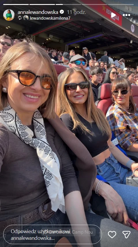 Trzy kobiety w okularach przeciwsłonecznych siedzące na stadionowych trybunach, uśmiechnięte, w otoczeniu tłumu widzów i przy słonecznej pogodzie.