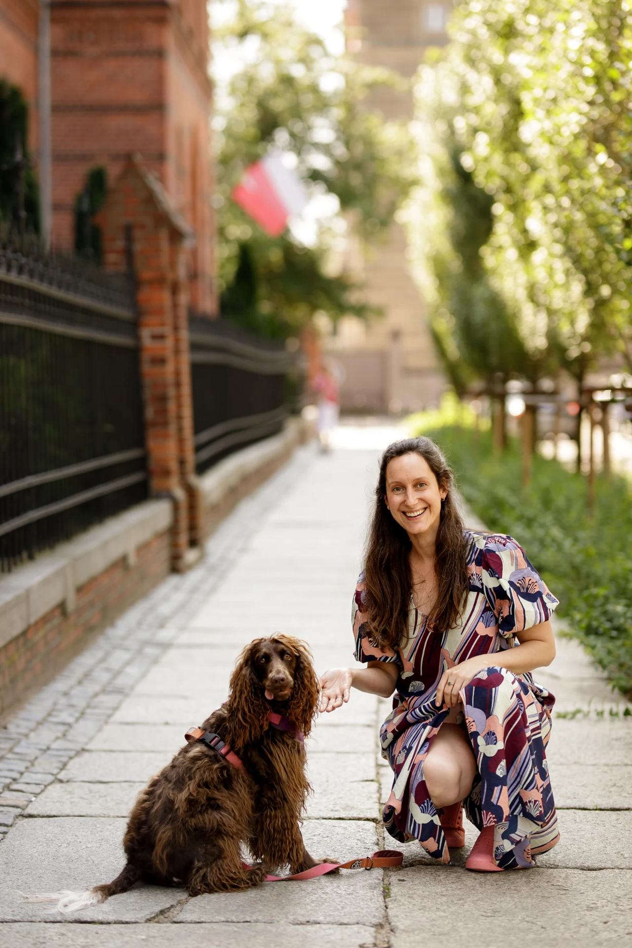 kobieta w kolorowej sukience klęczy na chodniku i trzyma za łapę brązowego psa z długą sierścią, oboje uśmiechnięci, w tle widoczne są drzewa oraz fragment metalowego ogrodzenia i polska flaga