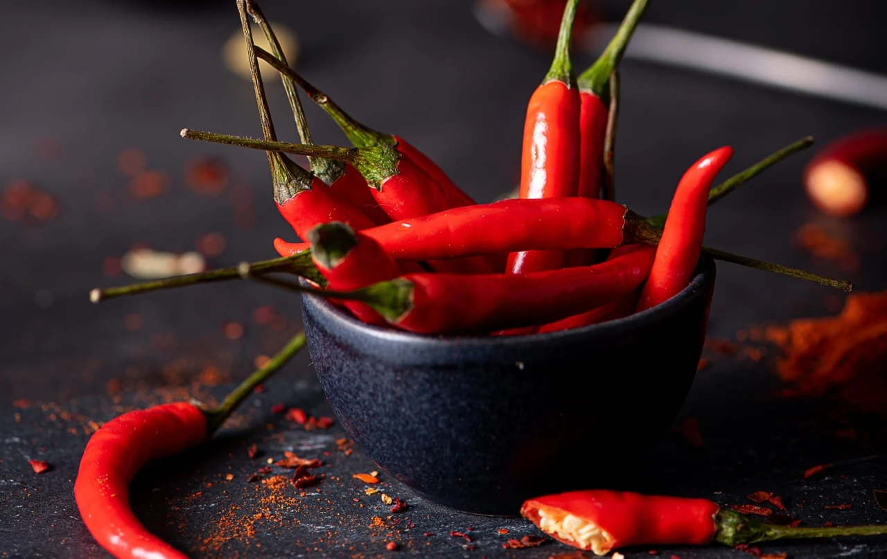 Czy papryczka chili przyspiesza metabolizm? Ekspertka rozwiewa wątpliwości Świeże czerwone papryczki chili w ciemnoniebieskiej miseczce na tle rozrzuconych przypraw i kawałków papryczek