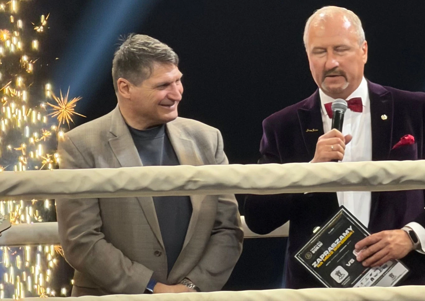 Dwóch elegancko ubranych mężczyzn stoi w ringu bokserskim; jeden trzyma mikrofon i mówi, a drugi spogląda na niego z uśmiechem; w tle widać sztuczne ognie.