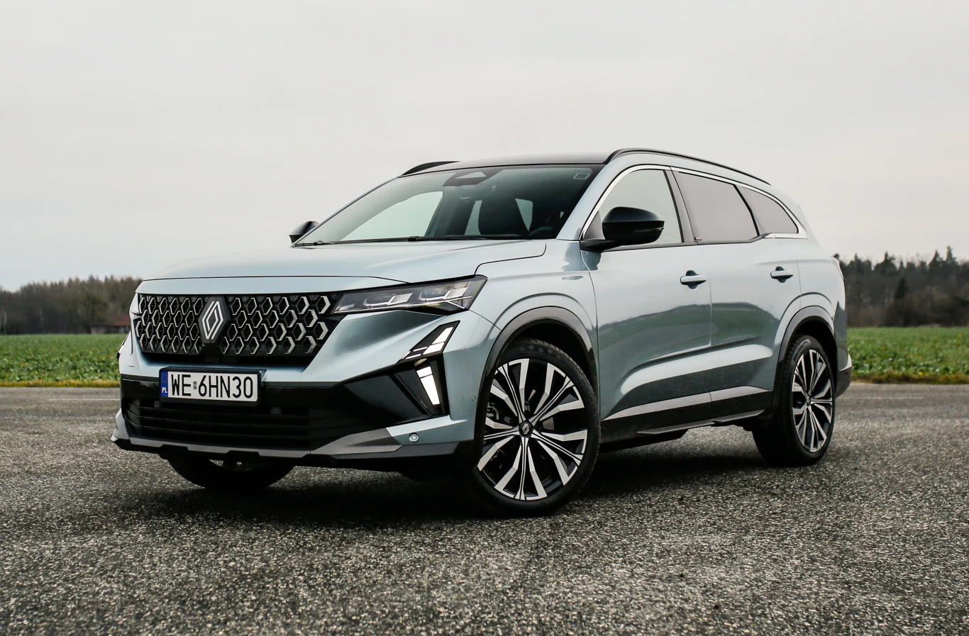 nowoczesny, srebrny SUV o dynamicznej sylwetce, masywnej osłonie chłodnicy z chromowanymi detalami, dużych aluminiowych felgach i smukłych reflektorach LED, zaparkowany na otwartej przestrzeni z polami i drzewami w tle