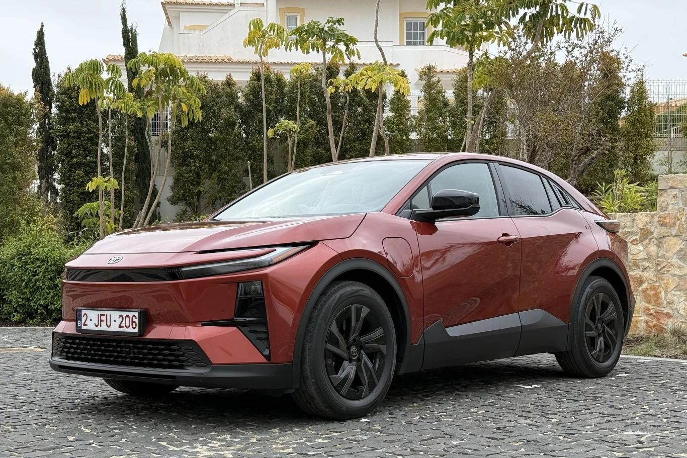 czerwony nowoczesny SUV o dynamicznej sylwetce, czarnych felgach i stylowych, ostrych liniach na karoserii, zaparkowany na brukowanym podjeździe w otoczeniu zielonych drzew i kamiennego muru