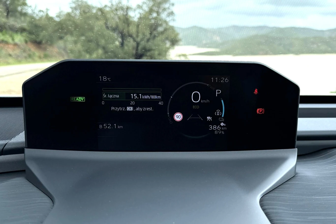 cyfrowy wyświetlacz kokpitu samochodu elektrycznego pokazujący aktualną prędkość, zasięg 386 km, poziom naładowania baterii 89% oraz zużycie energii 15,1 kWh/100 km, z widocznym otoczeniem drogi i zielenią za szybą