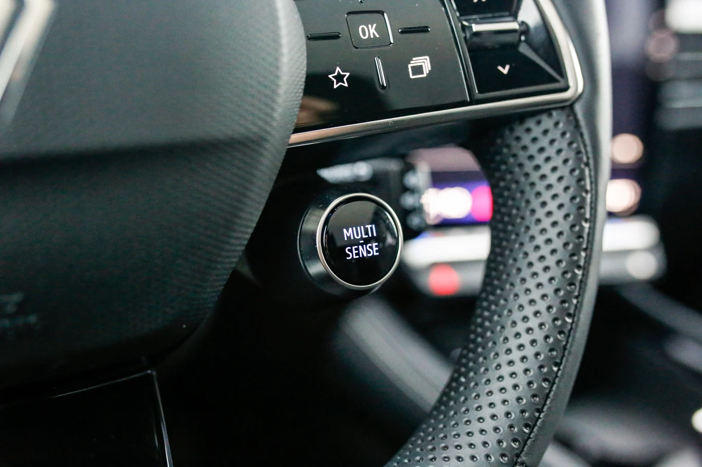 przycisk MULTI-SENSE umieszczony na perforowanej kierownicy samochodu, w tle wyraźnie widoczny panel sterowania z cyfrowymi przyciskami
