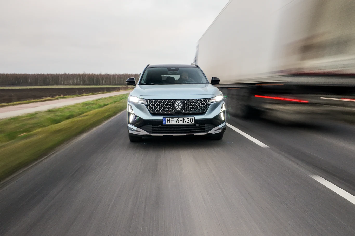 Srebrny SUV z charakterystycznym, dużym grillem oraz nowoczesnymi reflektorami jedzie drogą, mijając z dużą prędkością biały samochód ciężarowy po prawej stronie.