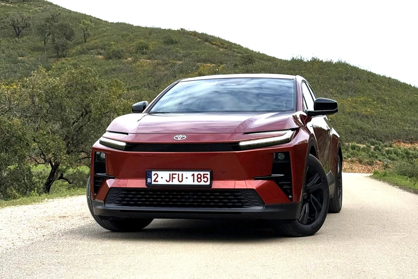 czerwony samochód marki Toyota o nowoczesnym, aerodynamicznym kształcie stoi na pustej asfaltowej drodze, otoczony zielonymi wzgórzami i drzewami, tablica rejestracyjna z belgijskim numerem