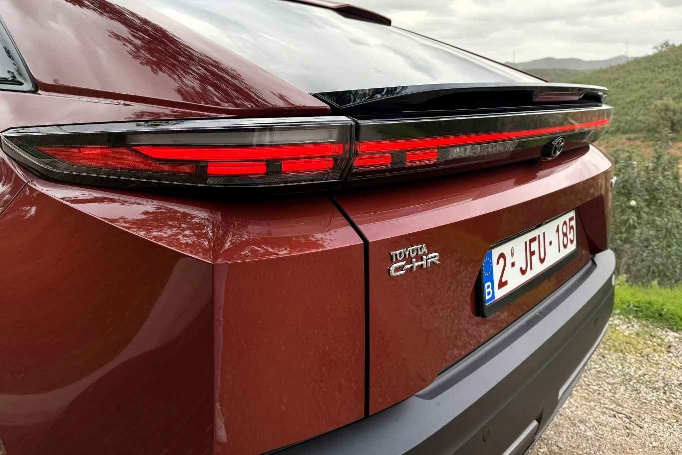 tylna część czerwonej Toyoty C-HR z nowoczesnym, poziomym pasem świateł LED, czarnym spoilerem oraz belgijską tablicą rejestracyjną na tle zielonego krajobrazu