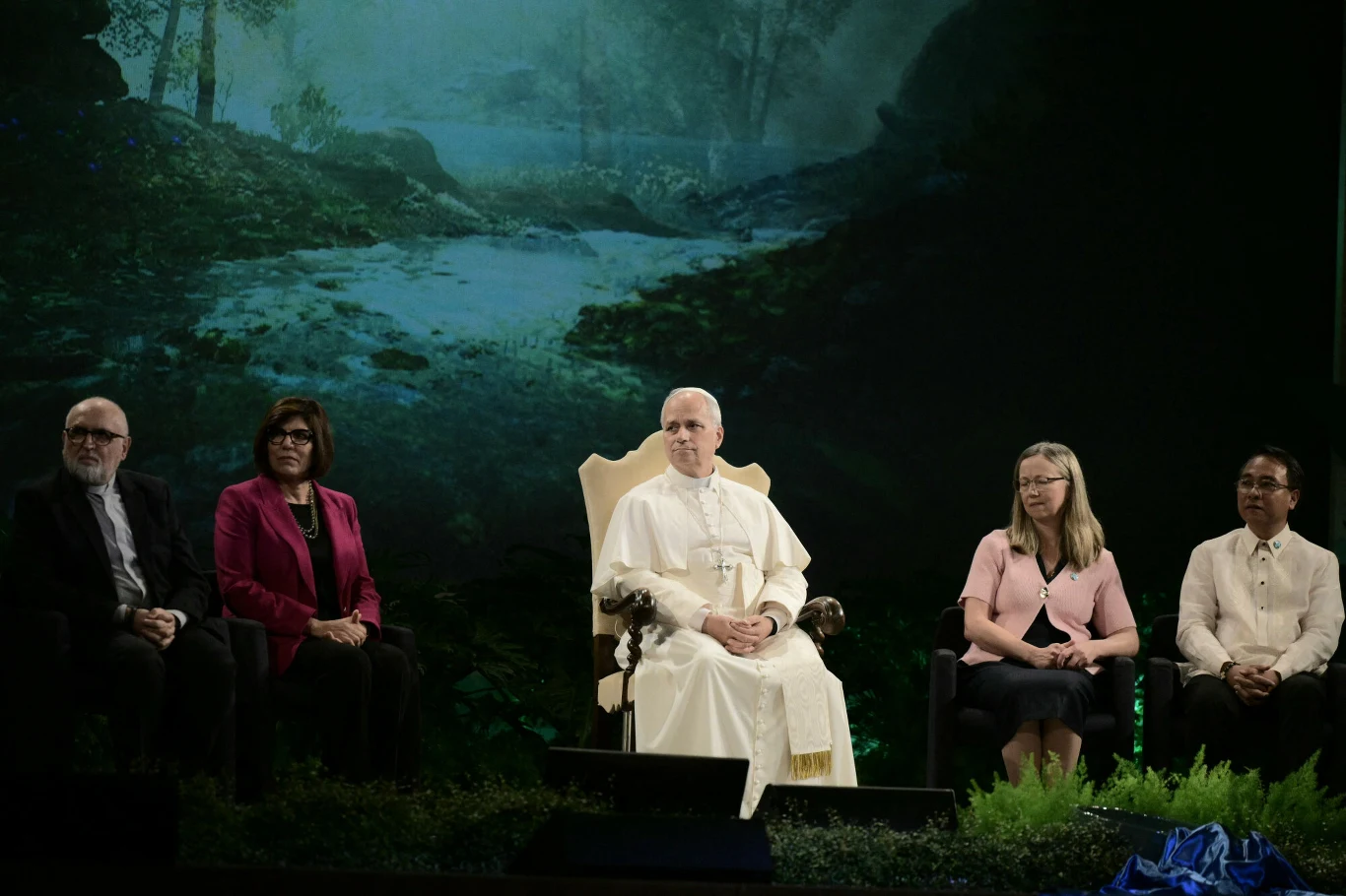 Pięć osób siedzących w jednym rzędzie na scenie, pośrodku osoba ubrana na biało na ozdobnym fotelu, po bokach dwie kobiety i dwóch mężczyzn w formalnych strojach, w tle nastrojowa leśna sceneria.