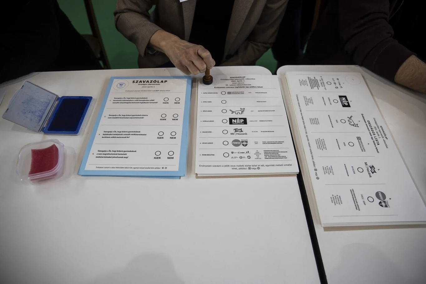Ręka osoby przybija pieczątkę na arkuszu wyborczym leżącym na stole, obok którego znajdują się podkładki, tusz oraz inne formularze do głosowania.
