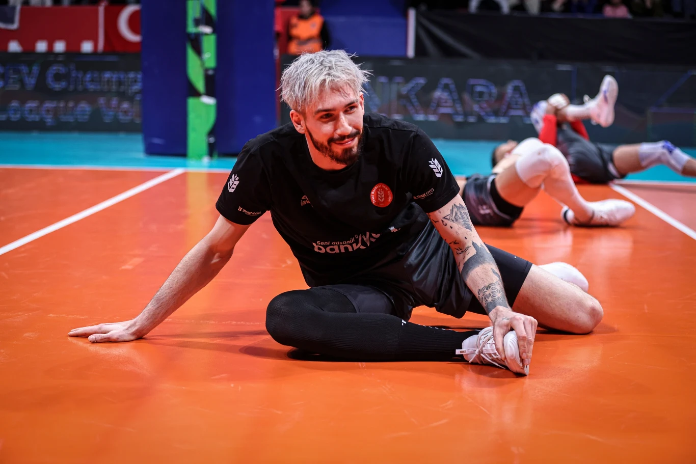 siatkarz z jasnymi włosami i tatuażem na lewym ramieniu wykonuje rozciągające ćwiczenie na parkiecie hali sportowej, w tle inny zawodnik również rozciąga się na podłodze