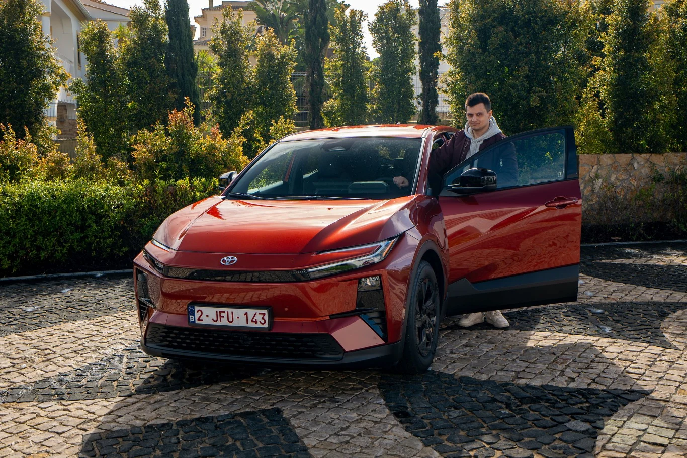 czerwony samochód marki Toyota z nowoczesnym, opływowym designem stoi zaparkowany na brukowanym podjeździe, a mężczyzna w jasnych ubraniach stoi przy otwartych drzwiach kierowcy
