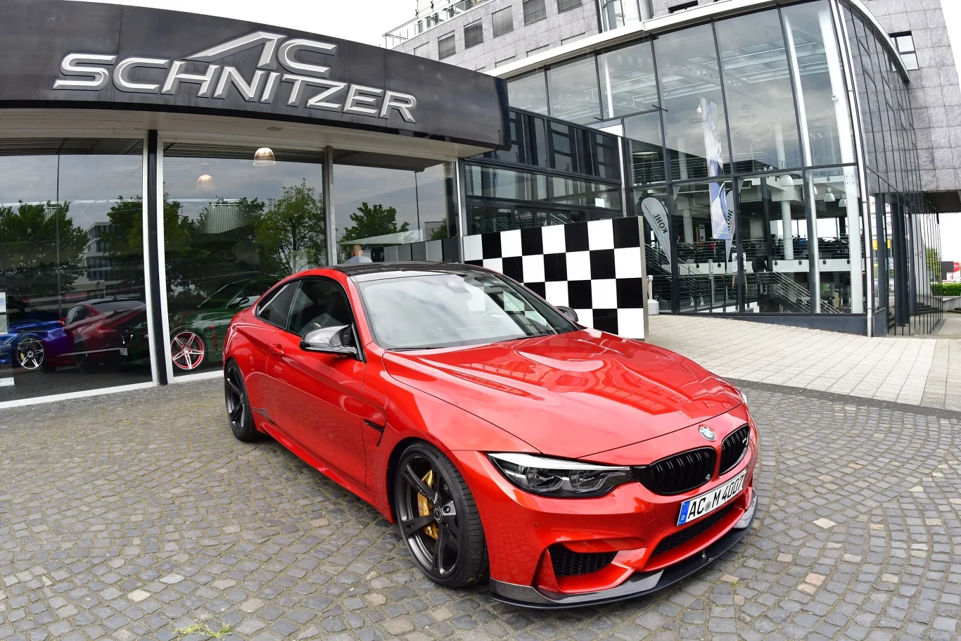 Czerwone sportowe BMW M4 z pakietem tuningowym AC Schnitzer stoi zaparkowane na bruku przed nowoczesnym budynkiem z przeszkloną fasadą oraz charakterystycznym szyldem tunera samochodowego.