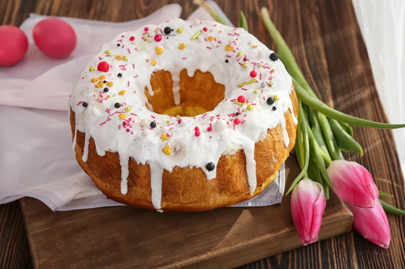 babka z białym lukrem polewana, ozdobiona kolorową posypką i jagodami, leżąca na drewnianej desce obok różowych tulipanów oraz dwóch różowych pisanek