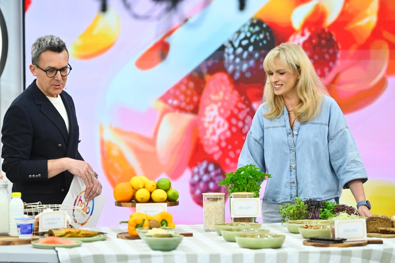 Maciej Kurzajewski, Justyna Mizera Dwoje osób stoi przy stole, na którym ułożone są świeże warzywa, owoce, zioła, nabiał i zdrowe produkty spożywcze, a w tle widoczna jest kolorowa grafika z owocami.