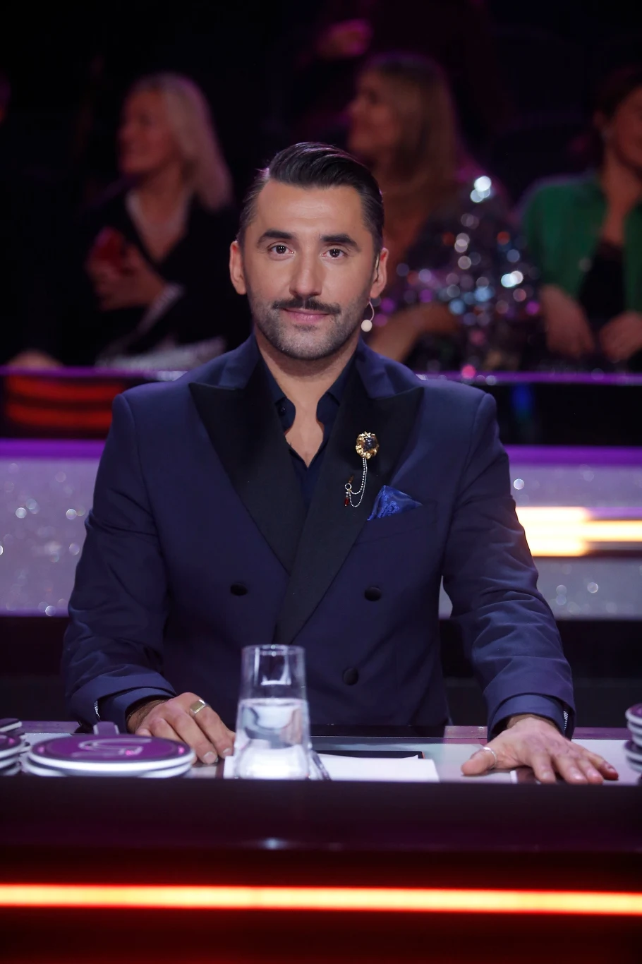 Mężczyzna w eleganckim, granatowym garniturze z przypinką siedzi przy stole jurorskim, patrząc wprost przed siebie, na stole szklanka z wodą oraz notatki, w tle rozmyta publiczność.