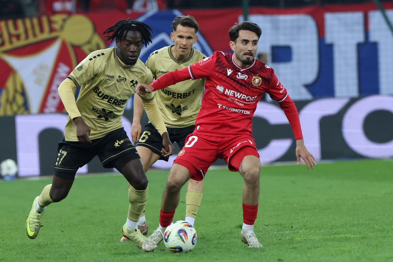 piłkarz w czerwonym stroju drybluje z piłką, mocno naciskany przez dwóch rywali w złotych koszulkach na tle stadionowych trybun wypełnionych kibicami