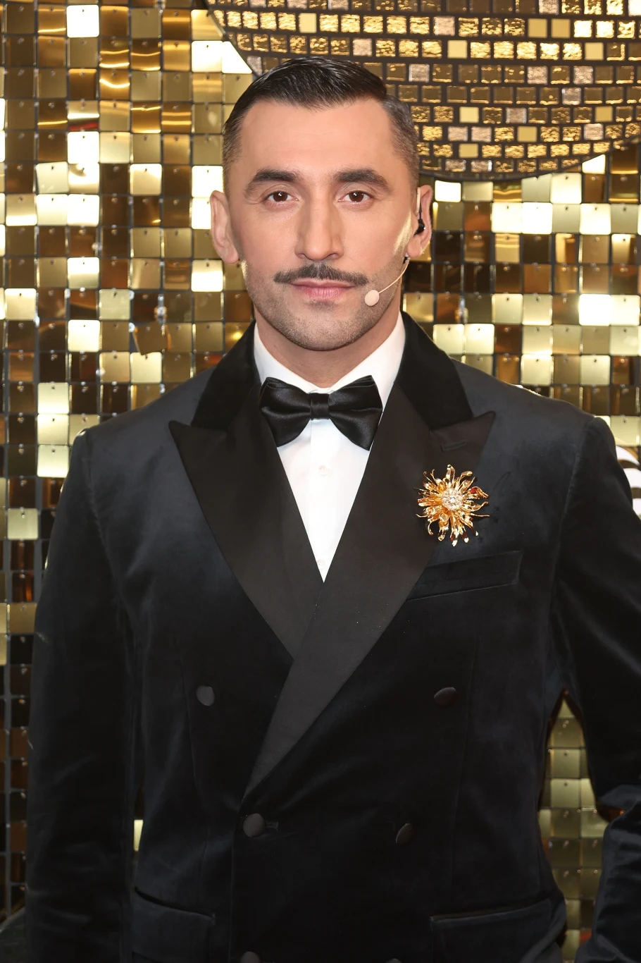 Elegancko ubrany mężczyzna w czarnym, aksamitnym smokingu z muchą i ozdobną broszką w kształcie gwiazdy na klapie, stojący na tle złotej, błyszczącej mozaiki.