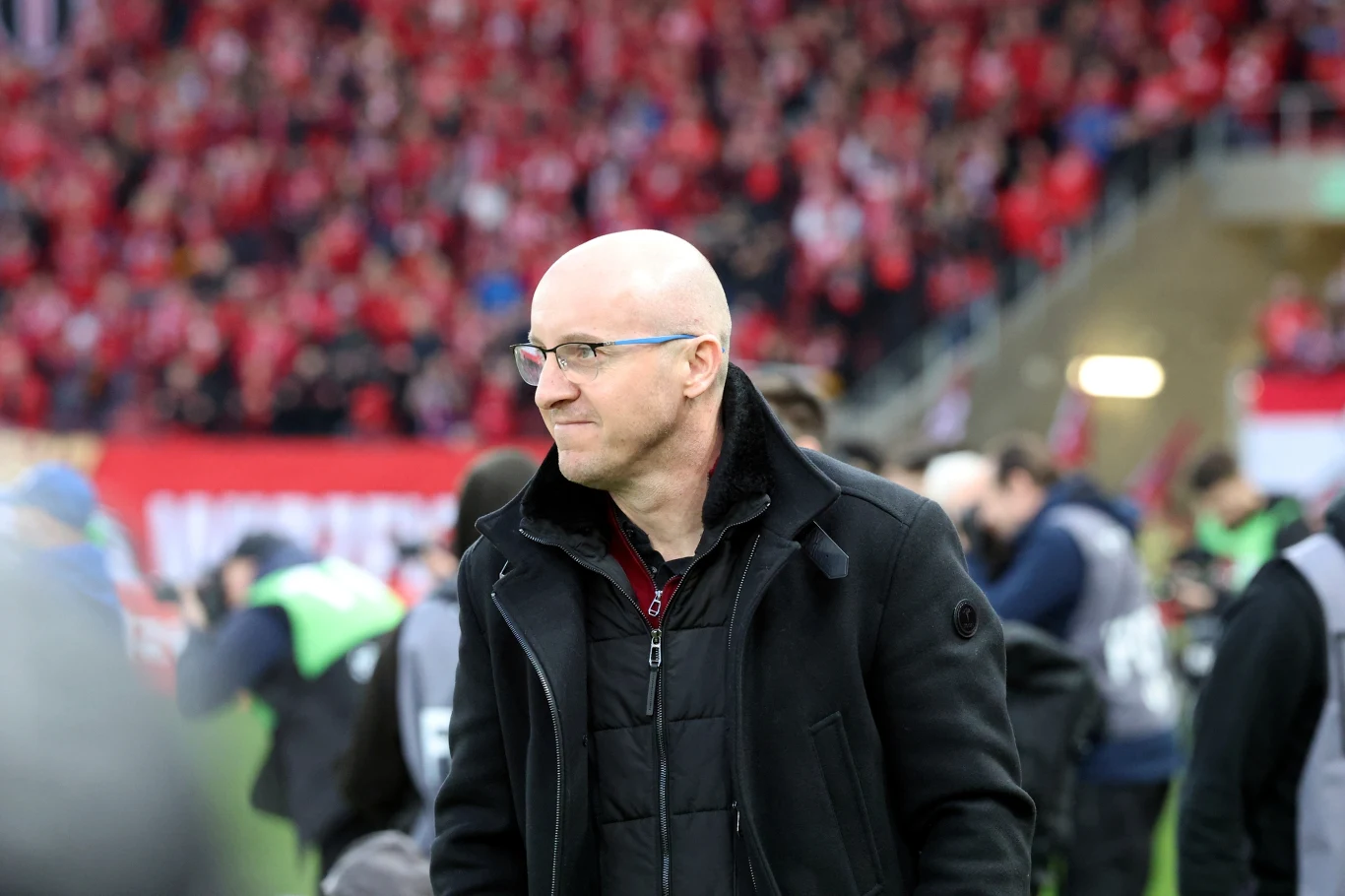 łysy mężczyzna w okularach i czarnej kurtce stoi na tle stadionu wypełnionego kibicami ubranymi na czerwono