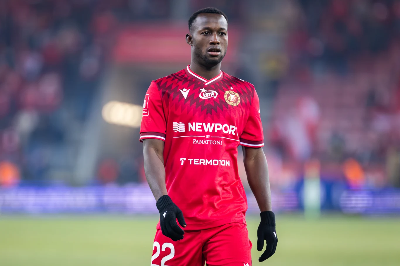 piłkarz w czerwonym stroju z numerem 22 i logotypami sponsorów spaceruje po murawie boiska podczas meczu, w tle rozmyta trybuna z widownią