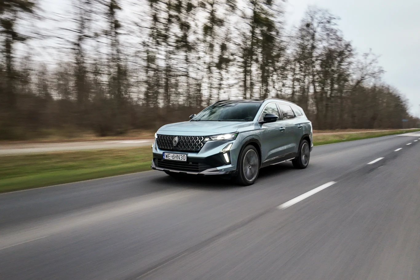 nowoczesny SUV w srebrnoszarym kolorze jedzie dynamicznie pustą asfaltową drogą, w tle rozmazany pas trawy i szpaler bezlistnych drzew