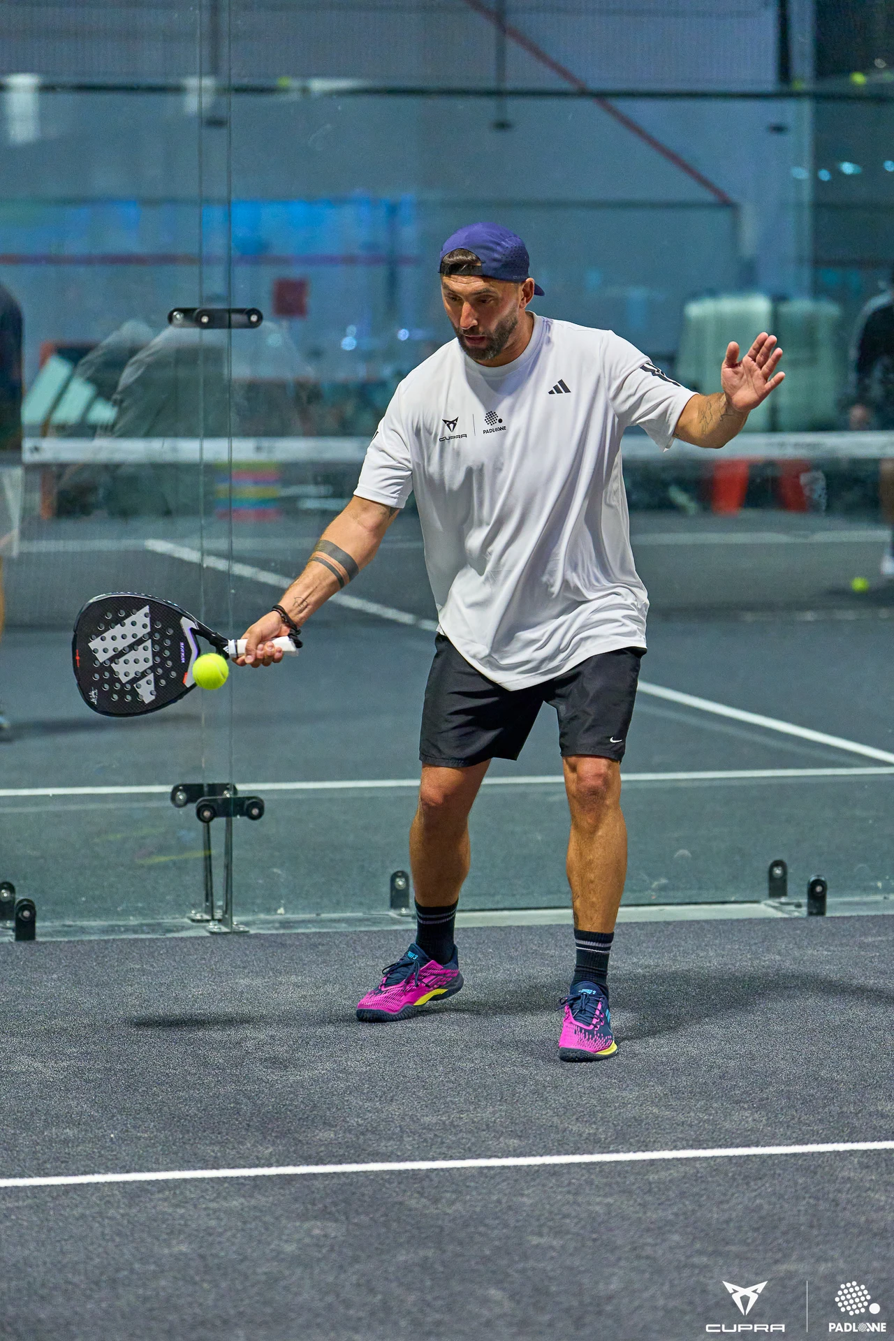 mężczyzna w sportowym stroju gra w padla, trzymając rakietę i przygotowując się do odbicia piłki na szklanym korcie