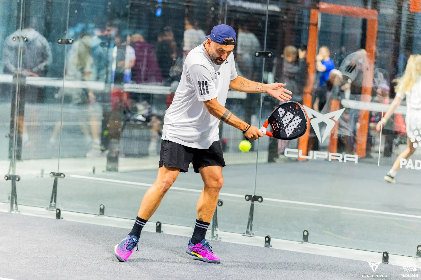 mężczyzna w sportowej odzieży gra w padla na korcie, wykonując uderzenie rakietą w kierunku żółtej piłki, wokół szklane ściany i widoczne odbicia postaci w tle