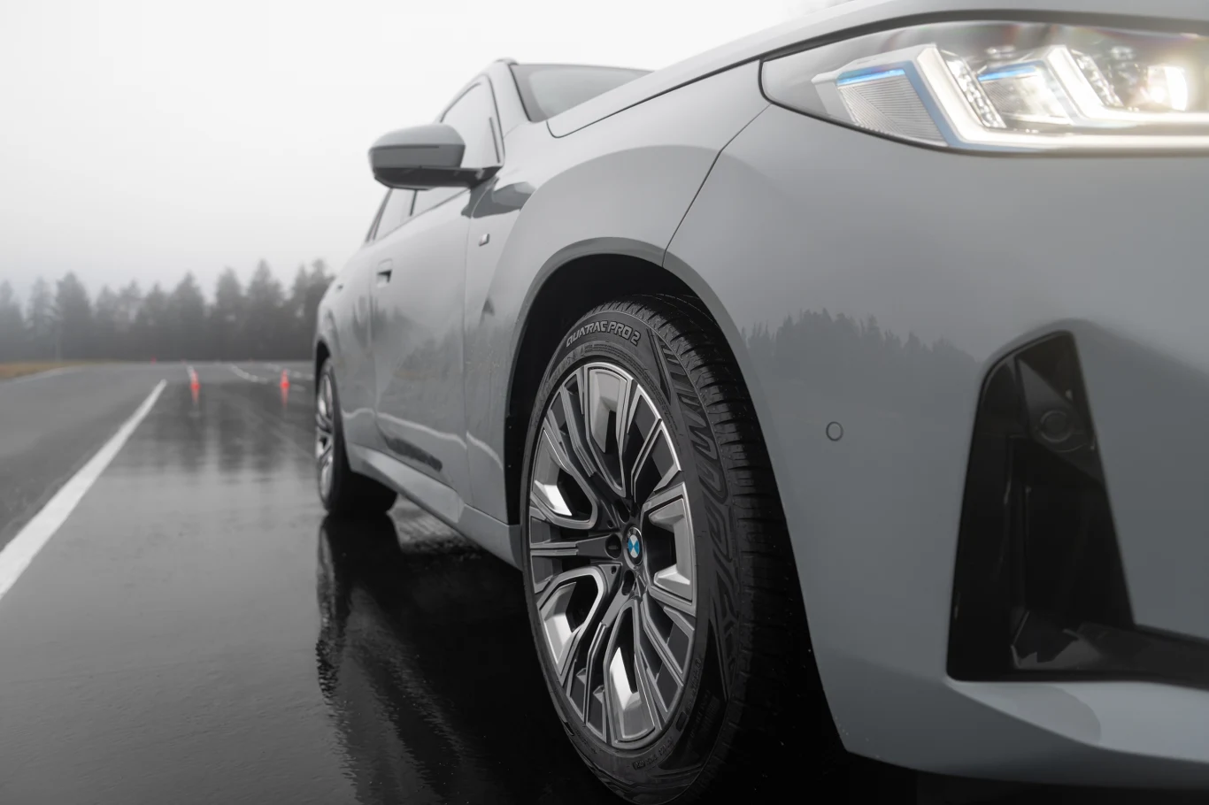Srebrny samochód BMW ustawiony na mokrej nawierzchni drogi, w tle widoczny rząd drzew oraz pomarańczowe pachołki na jezdni.