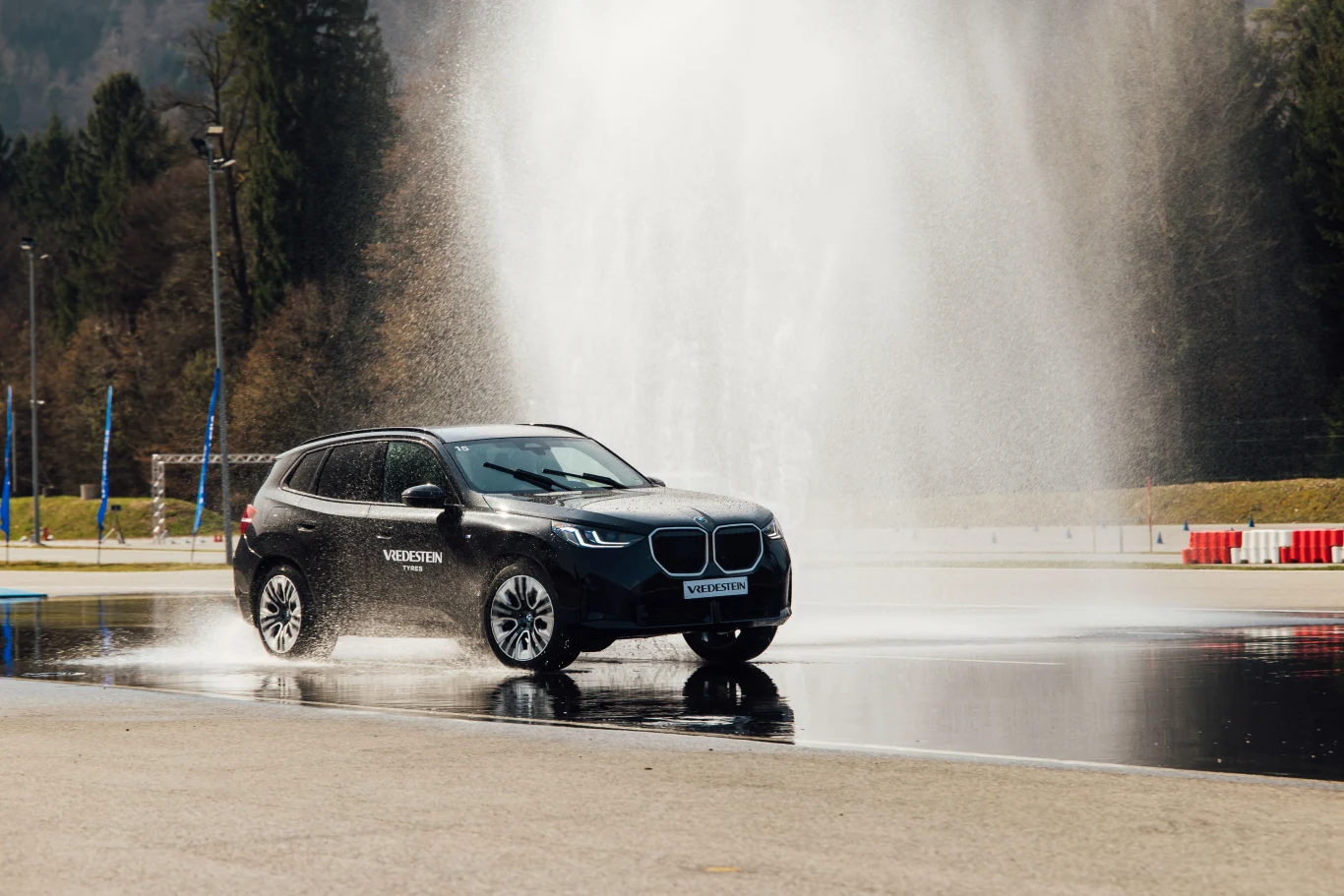 Czarny SUV marki BMW jedzie po mokrej nawierzchni, obok wysokiego strumienia wody tryskającego z ziemi, odbijając się w powierzchni drogi; wokół widoczne są drzewa i barierki toru testowego.
