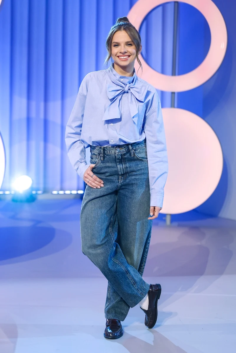 uśmiechnięta kobieta ubrana w jasnoniebieską koszulę z dużą kokardą przy szyi oraz szerokie dżinsy, stojąca pewnie na tle dekoracyjnych, abstrakcyjnych okręgów i niebieskich zasłon