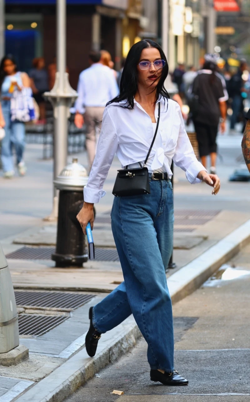 kobieta w białej koszuli i niebieskich dżinsach idzie chodnikiem po mieście, trzymając w ręce książkę, z czarną torebką przewieszoną przez ramię, ma długie ciemne włosy i okulary, wokół ruch uliczny i przechodnie w tle