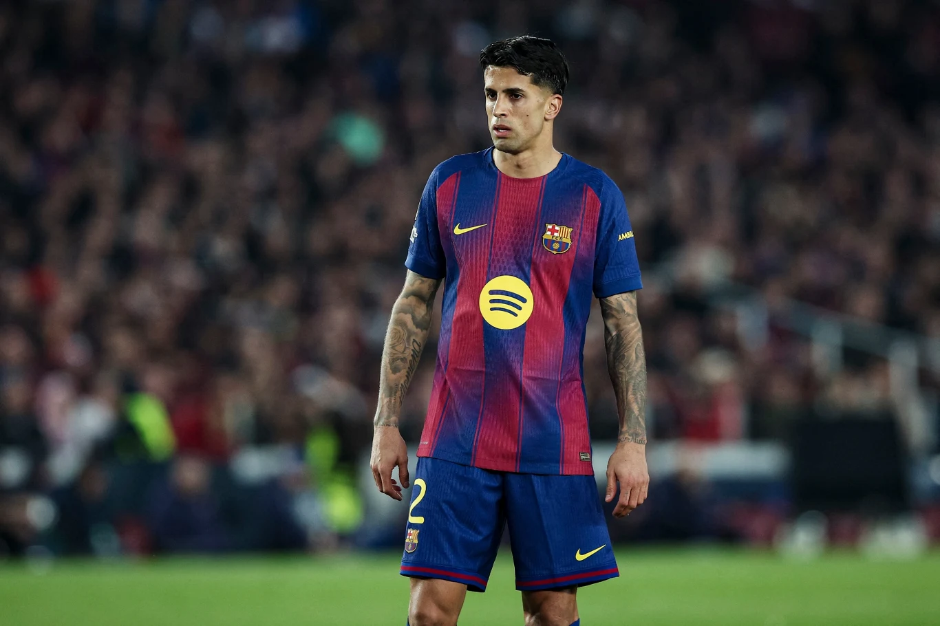 piłkarz w niebiesko-granatowej koszulce FC Barcelona z żółtym numerem 2 oraz herbem klubu na piersi stoi na murawie podczas meczu, w tle rozmyty tłum kibiców na trybunach