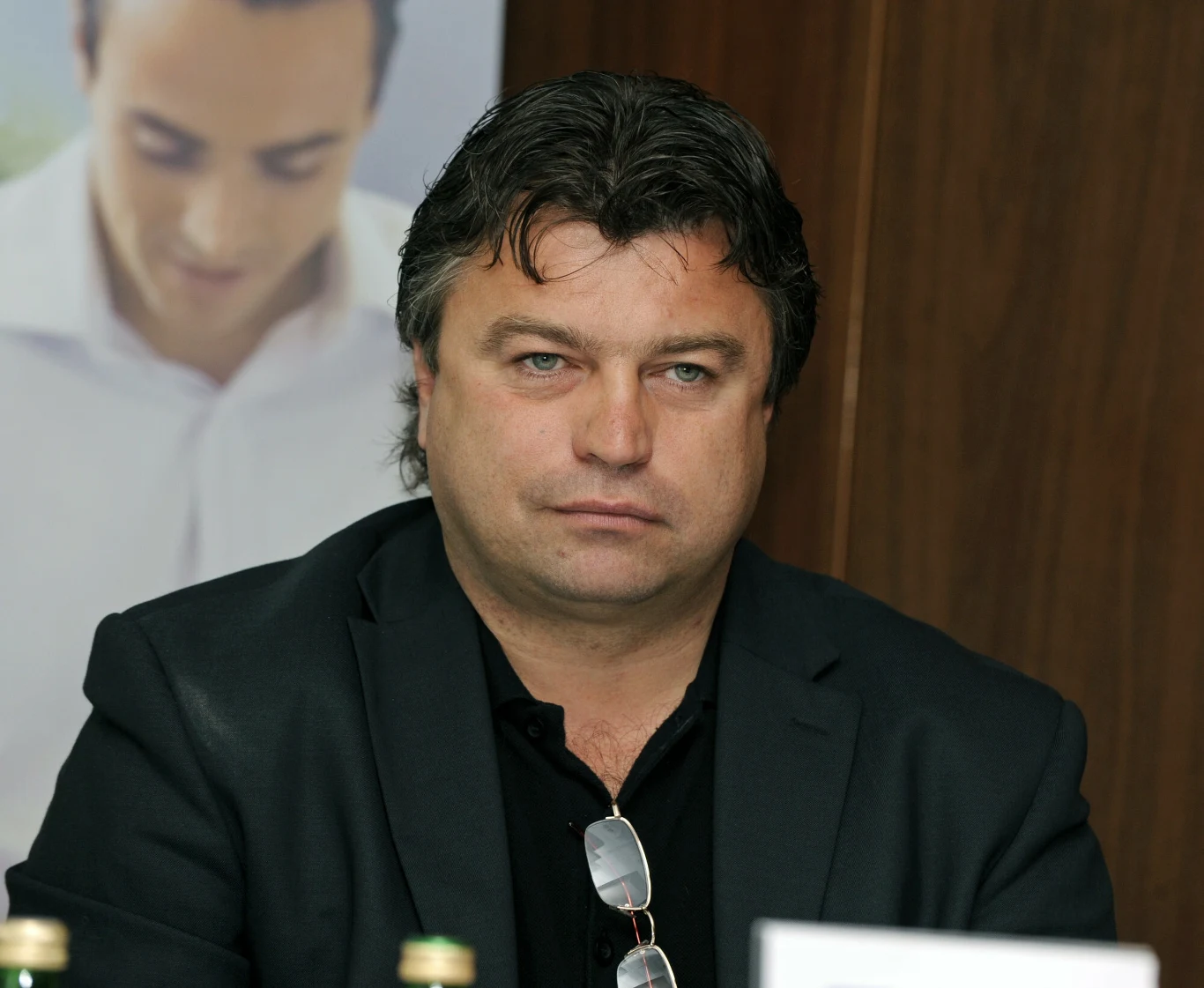 mężczyzna w średnim wieku o ciemnych włosach, ubrany w czarną marynarkę i koszulę, siedzi przy stole z poważnym wyrazem twarzy; na szyi zawieszone okulary, w tle fragment plakatu z wizerunkiem innej osoby