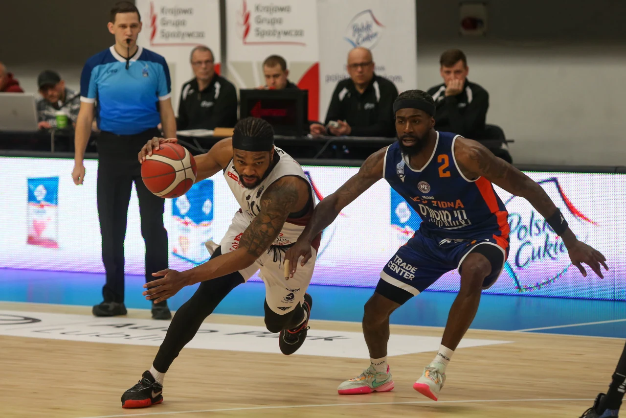 zawodnik w białym stroju walczy o piłkę podczas dynamicznej akcji na boisku do koszykówki, za nim znajduje się przeciwnik w niebieskim stroju próbujący go powstrzymać, w tle sędzia i osoby przy stoliku sędziowskim