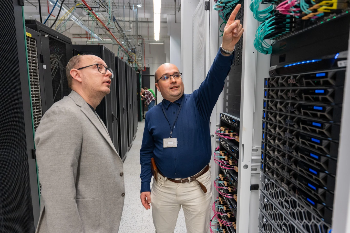 Dwóch mężczyzn w serwerowni, jeden z nich wskazuje na przewody sieciowe podłączone do serwera, obok znajdują się szafy rackowe z widocznymi podzespołami komputerowymi.