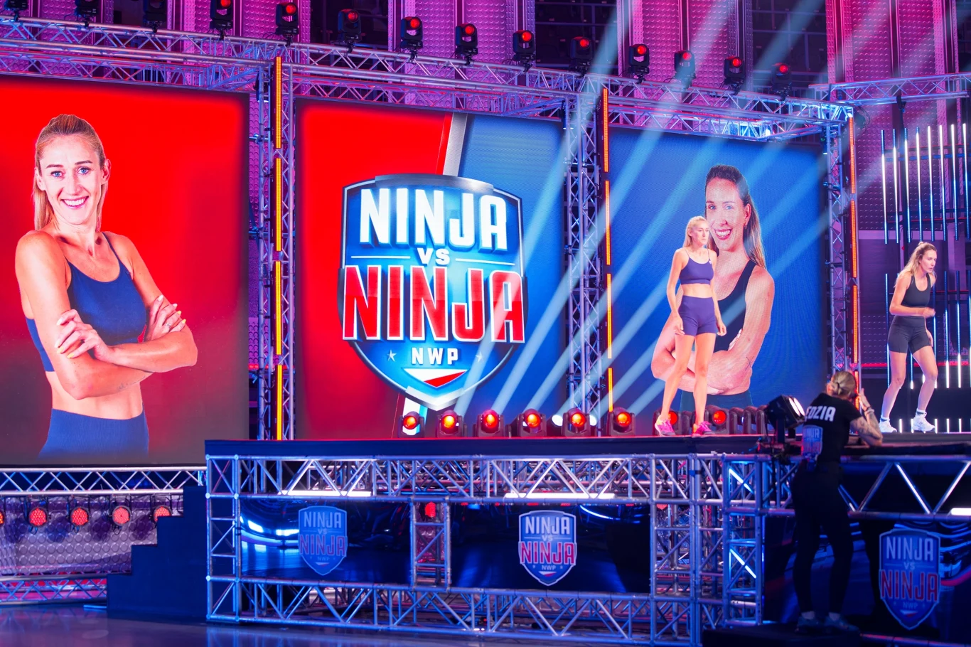 arena telewizyjnego show Ninja vs Ninja z zawodniczką w sportowym stroju stojącą na podium, wokół kolorowe światła, duże ekrany z logotypem programu i wizerunkiem zawodniczki, dynamiczna atmosfera sportowej rywalizacji