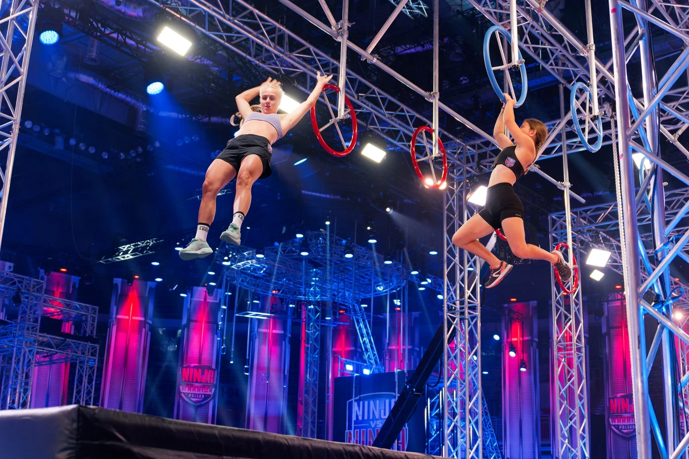 Dwie kobiety w sportowych strojach pokonują wiszące przeszkody na torze Ninja Warrior, jedna trzyma się czerwonego okręgu, druga jest w trakcie przeskoku między obręczami, na tle kolorowego oświetlenia i metalowej konstrukcji.