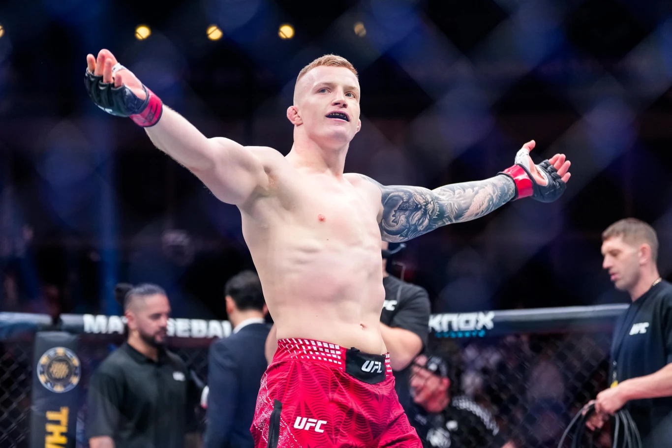 Zawodnik MMA stojący na środku oktagonu z uniesionymi rękami, ubrany w czerwone spodenki z logo UFC, wokół niego sędziowie i trenerzy w trakcie walki lub jej zakończenia.