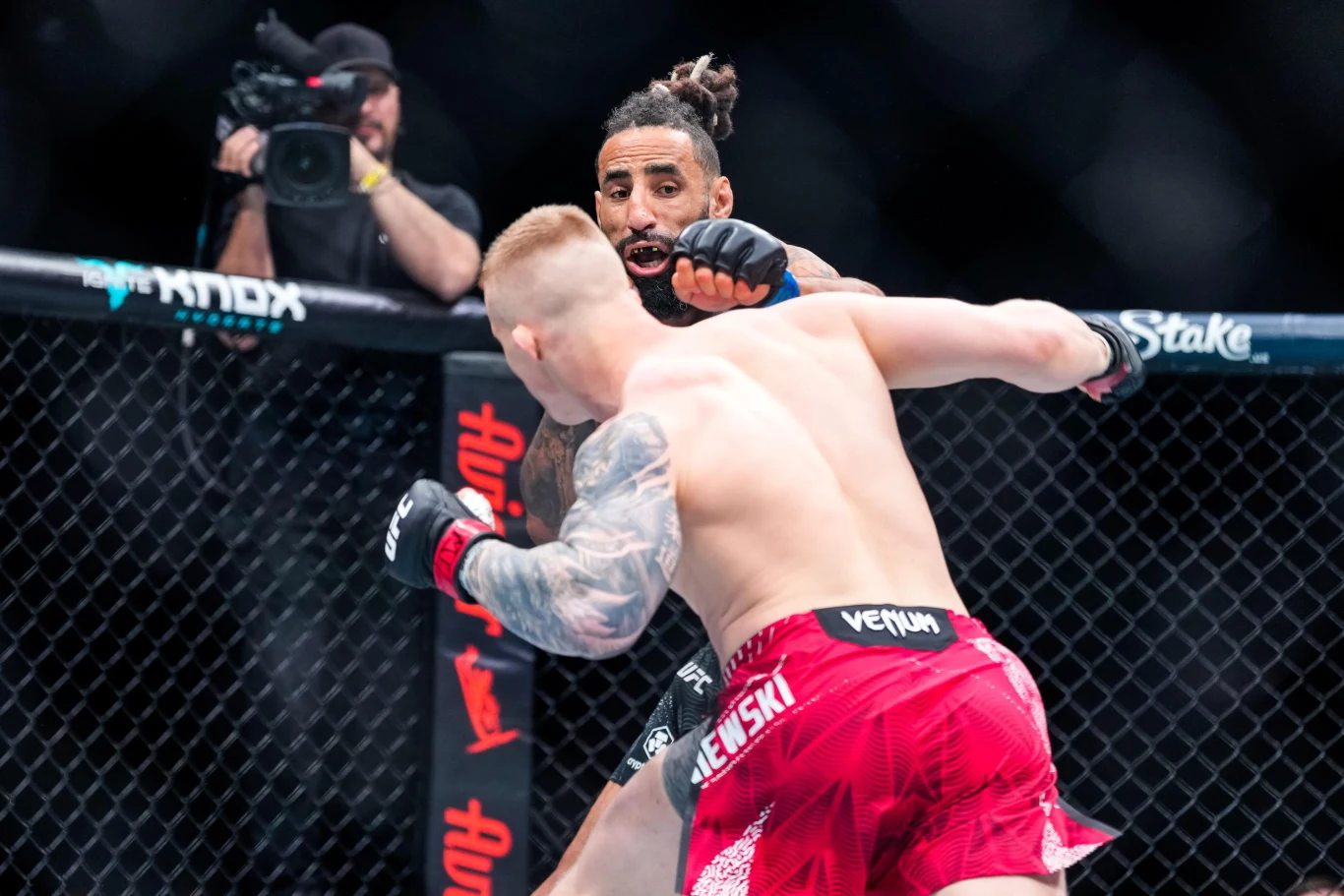 dwóch zawodników walczących w oktagonie MMA, jeden z nich uderza przeciwnika pięścią, w tle widoczny kamerzysta zapisujący przebieg walki