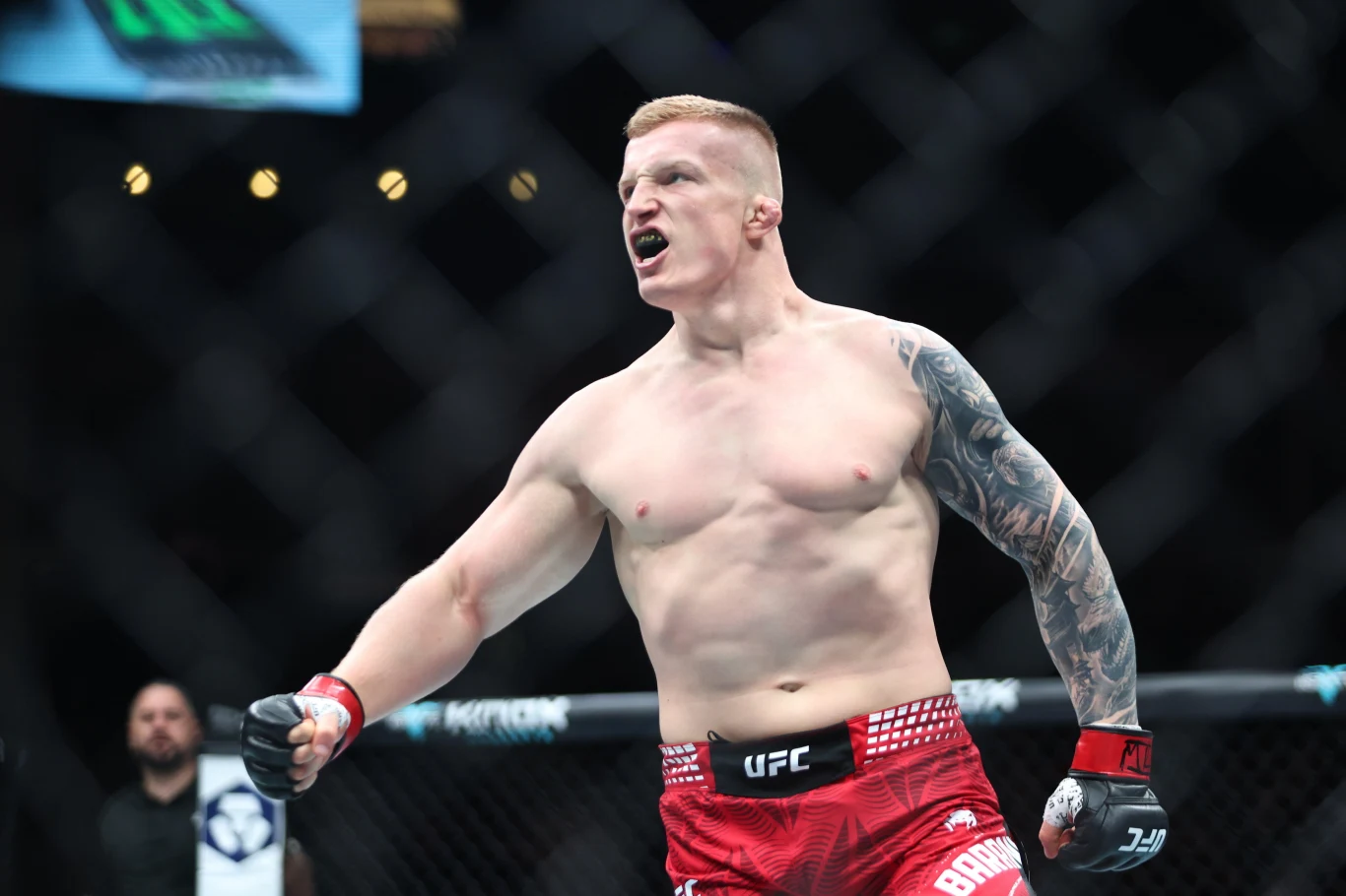 Zawodnik MMA w czerwonych spodenkach z uniesioną pięścią i wyrazem triumfu na twarzy, stojący w oktagonie UFC, z widocznym tatuażem na ręce.