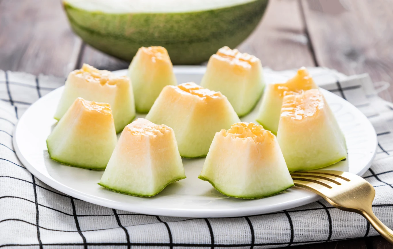 Kawałki świeżego melona ułożone na białym talerzu, na którym leży również złoty widelec, wszystko umieszczone na biało-czarnej ściereczce z kratką.