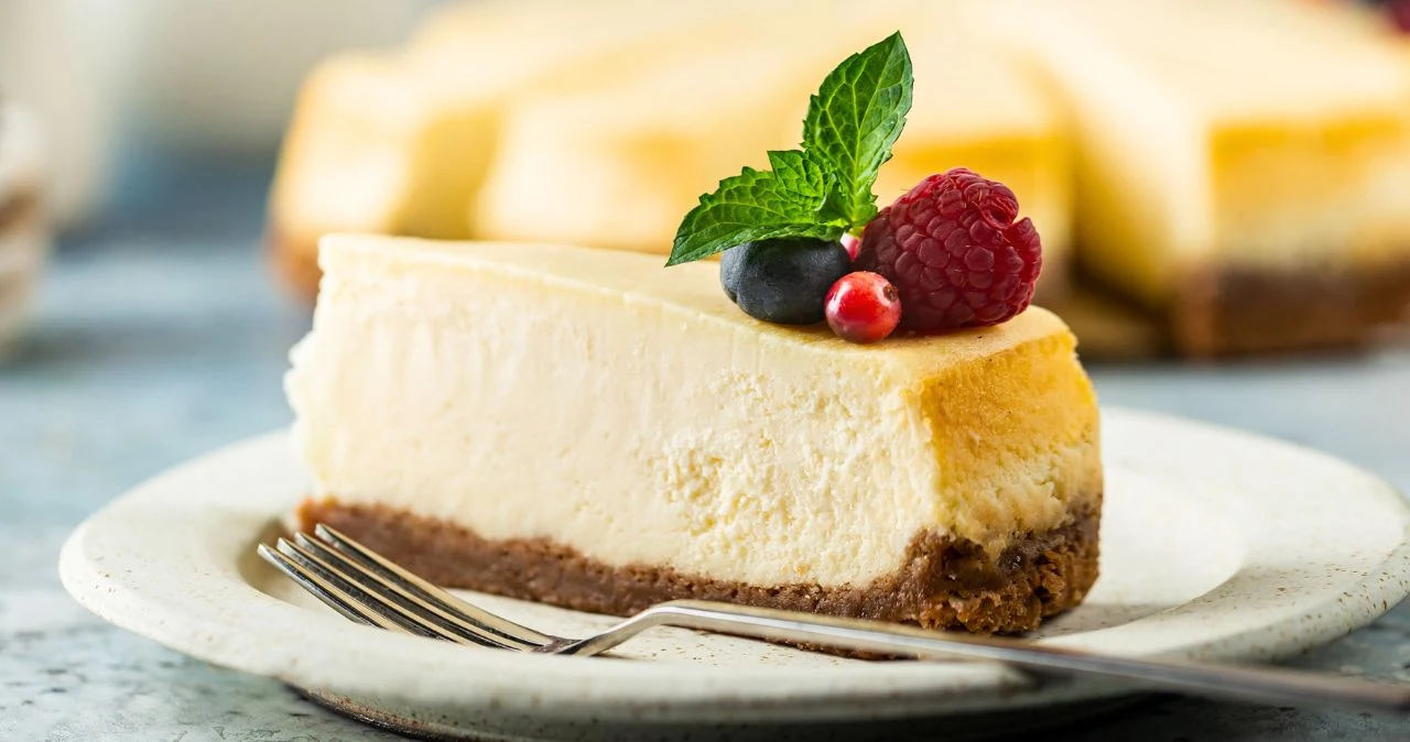 Kawałek kremowego sernika z kruchego spodu ozdobiony listkiem mięty oraz świeżymi owocami: maliną, borówką i porzeczką, podany na białym talerzu z widelcem.