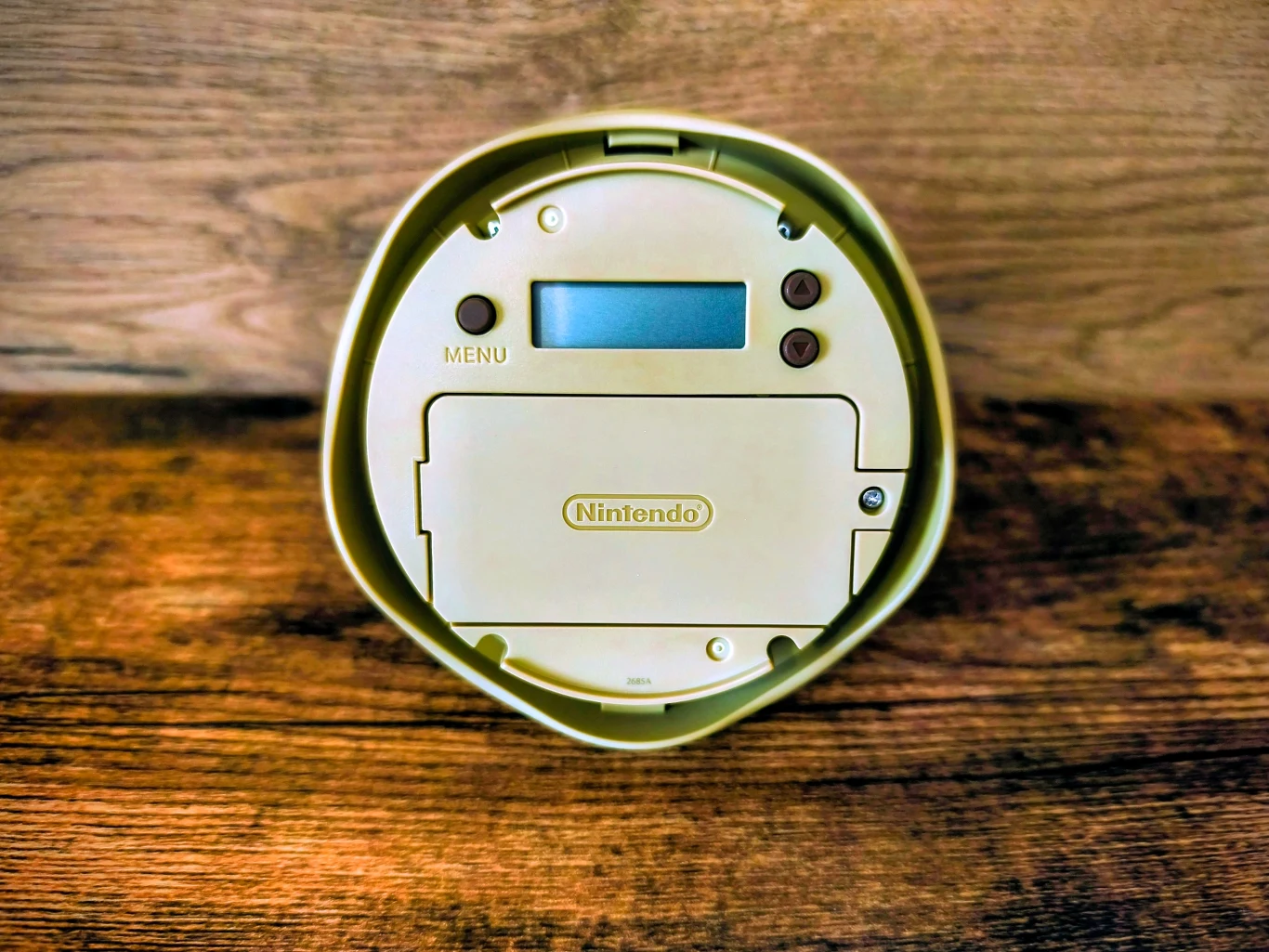 okrągłe elektroniczne urządzenie Nintendo z wyświetlaczem LCD, trzema przyciskami, oznaczeniem 'MENU' i logotypem Nintendo na obudowie, ustawione na drewnianej powierzchni