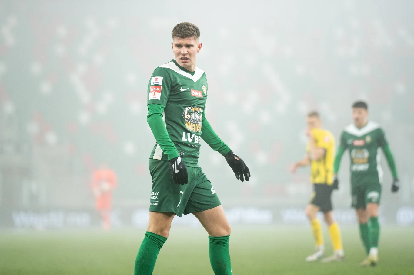 piłkarz w zielonym stroju podczas meczu na boisku, otoczony przez innych zawodników ubraną w zielone i żółte stroje, gra rozgrywa się w zamglonej atmosferze