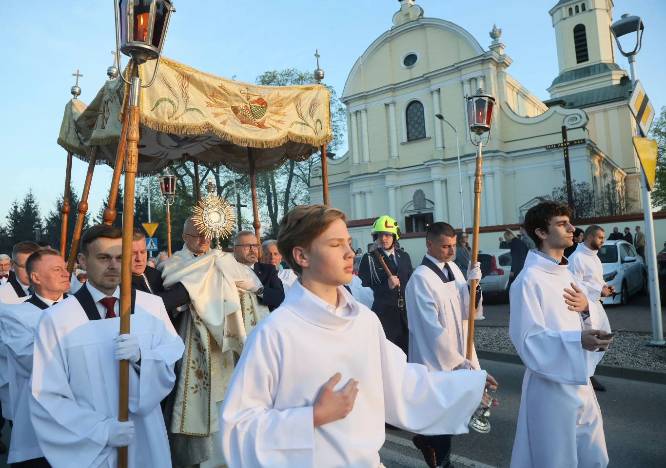 Procesja religijna prowadząca przez ulicę, kapłan niesie monstrancję pod baldachimem, otoczony przez ministrantów w białych szatach oraz wiernych; w tle widoczny kościół.