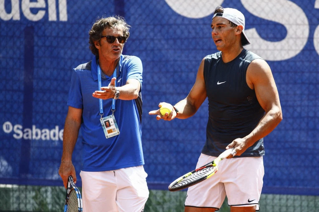 Dwóch mężczyzn na korcie tenisowym, jeden w koszulce na ramiączkach z rakietą i piłką, drugi w niebieskiej koszulce gestykulujący podczas rozmowy; za nimi widoczny baner reklamowy.