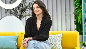 Maria Dębska na żółtej kanapie, ubrana w czarną bluzkę i jasnoniebieskie jeansy, z subtelnym uśmiechem na twarzy; za nią poduszki dekoracyjne, obok widoczne są kwiaty w doniczkach.