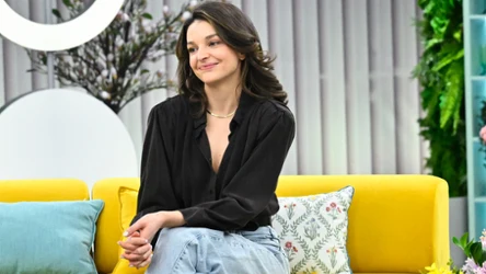 Maria Dębska na żółtej kanapie, ubrana w czarną bluzkę i jasnoniebieskie jeansy, z subtelnym uśmiechem na twarzy; za nią poduszki dekoracyjne, obok widoczne są kwiaty w doniczkach.